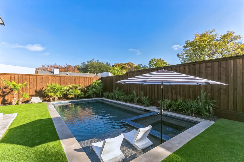 A rectangular swimming pool features two sheer descent water curtains flowing from a stone retaining wall. The pool is surrounded by a light-colored patio and a vibrant green lawn, with a tall wooden privacy fence and a large, leafy tree in the background under a soft, overcast sky.