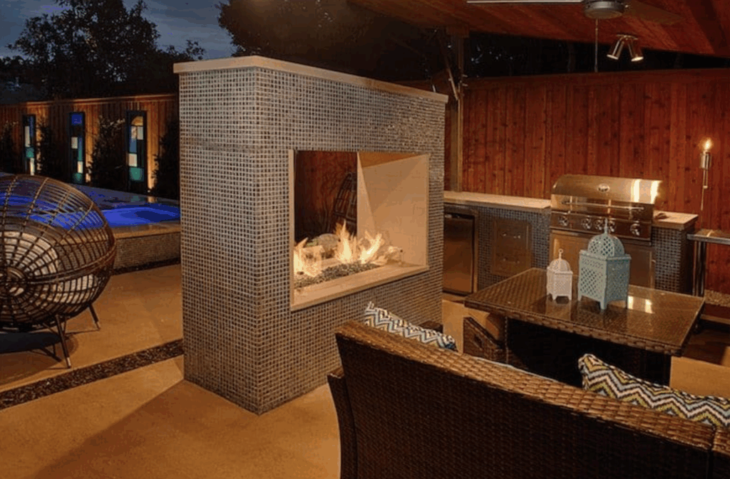An evening view of a luxurious outdoor living space featuring a double-sided gas fireplace covered in gray mosaic tiles. The area includes an outdoor kitchen with a stainless steel grill, a dining set with wicker furniture, and a glimpse of a glowing swimming pool in the background.