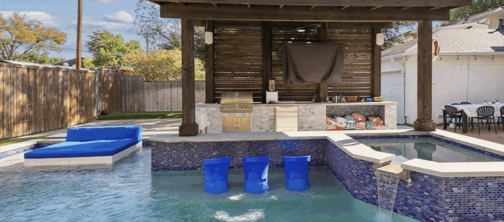 A modern backyard renovation showcasing a geometric pool with dark blue water and a tanning ledge under a striped umbrella. The pool is surrounded by vibrant green turf and stone steps leading to a raised patio. On the patio, a sleek black metal pergola with a translucent roof shades a white outdoor sectional sofa and a wall-mounted TV. The house features charcoal gray brick and board-and-batten siding, creating a cohesive, high-end aesthetic.