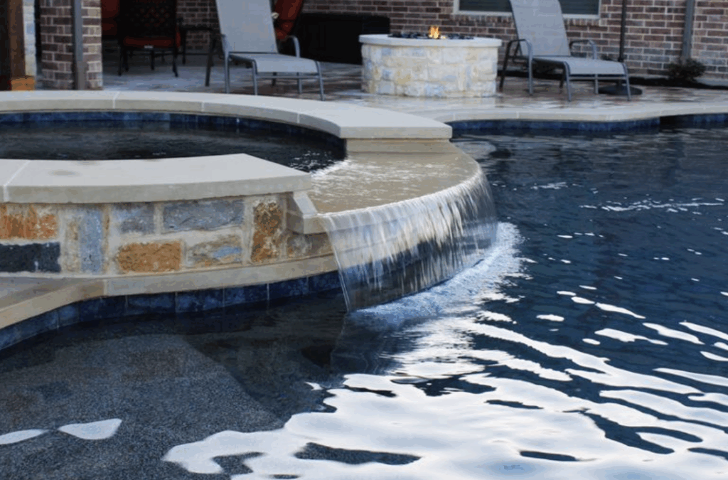 A close-up view of a custom stone hot tub overflowing into a dark-bottom swimming pool via a wide spillway waterfall. In the background, a stone fire pit and lounge chairs sit on a paved patio next to a brick house.