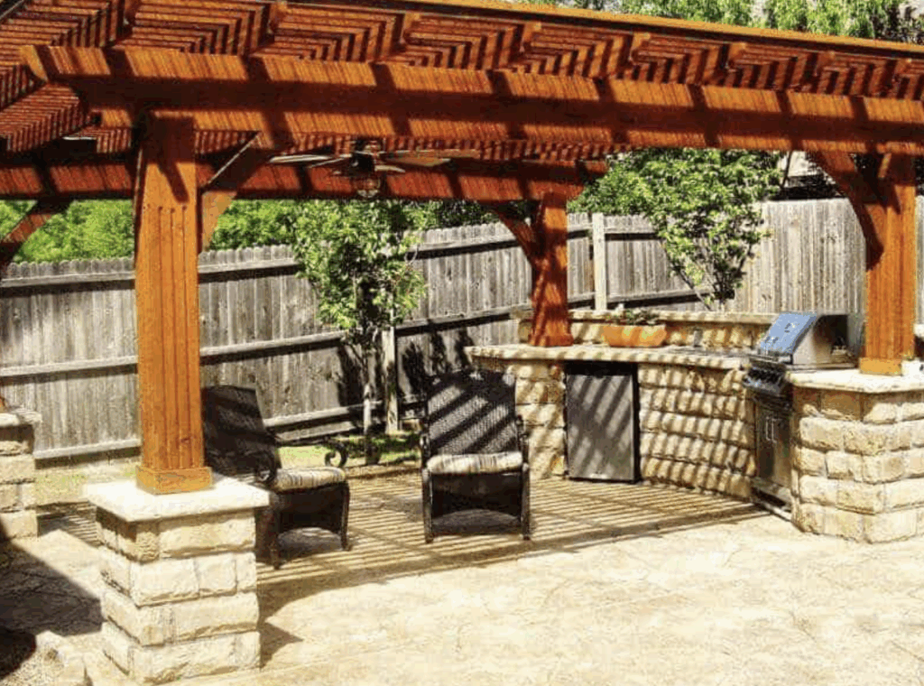 A rustic outdoor living area featuring a large wooden pergola with a ceiling fan, a stone-based outdoor kitchen with a grill and mini-fridge, and a seated patio area under a shaded lattice roof.