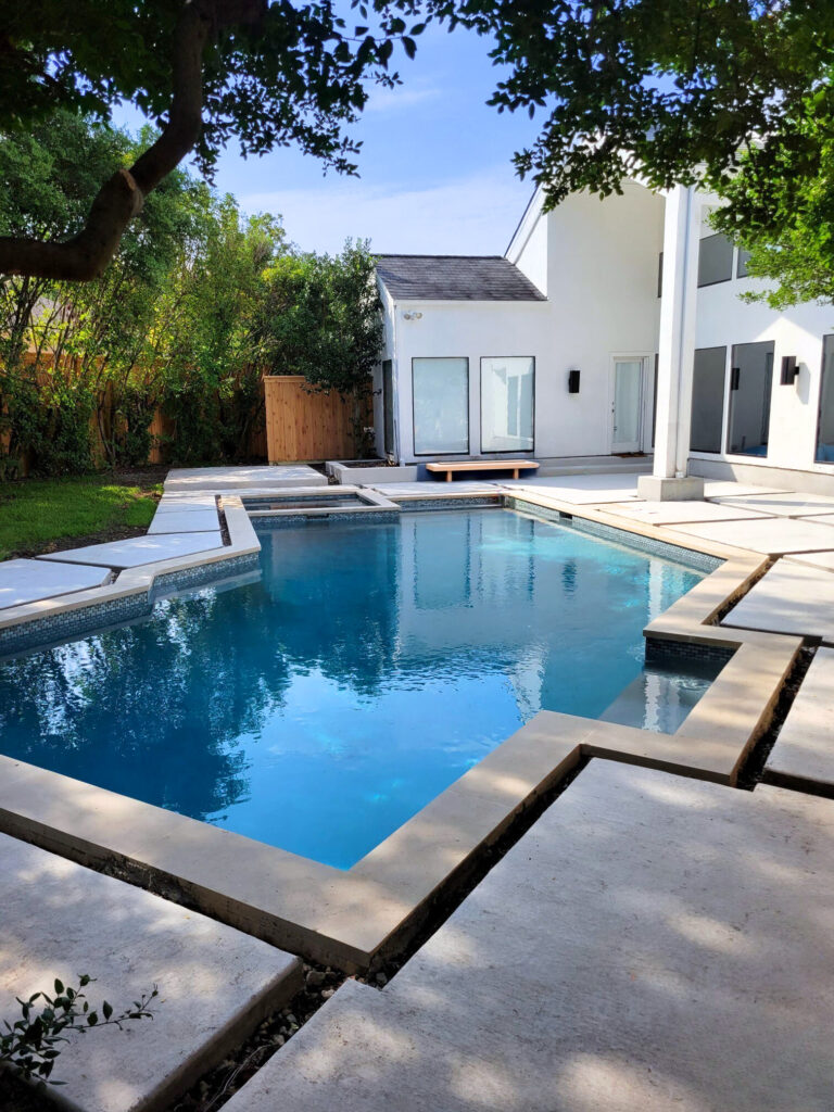 Modern backyard pool with geometric design, elevated spa, concrete deck, white minimalist house, and surrounding trees for privacy.