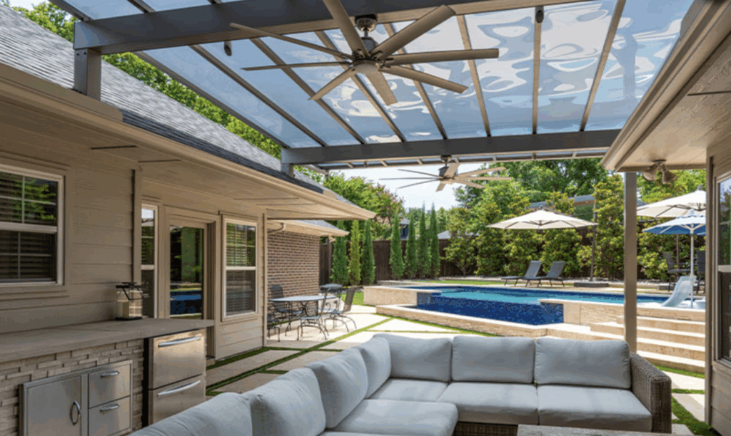 Modern backyard with covered patio, ceiling fans, L-shaped sofa, outdoor kitchen, raised pool with waterfall feature, and shaded lounge chairs.