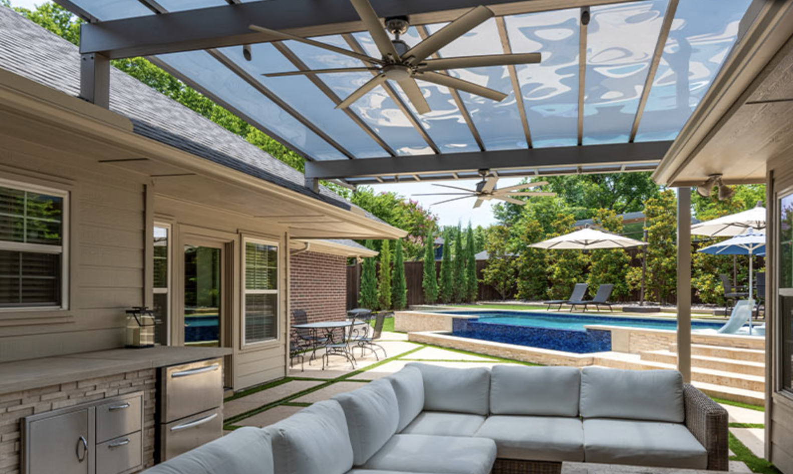 Modern backyard with covered patio, ceiling fans, L-shaped sofa, outdoor kitchen, raised pool with waterfall feature, and shaded lounge chairs.