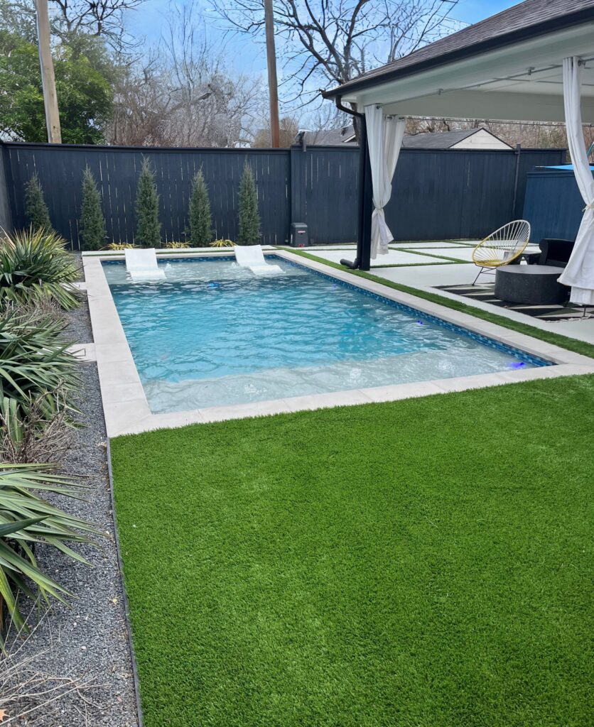Modern backyard pool with submerged lounge chairs, artificial grass, concrete pavers, and covered patio with fire pit and privacy landscaping.