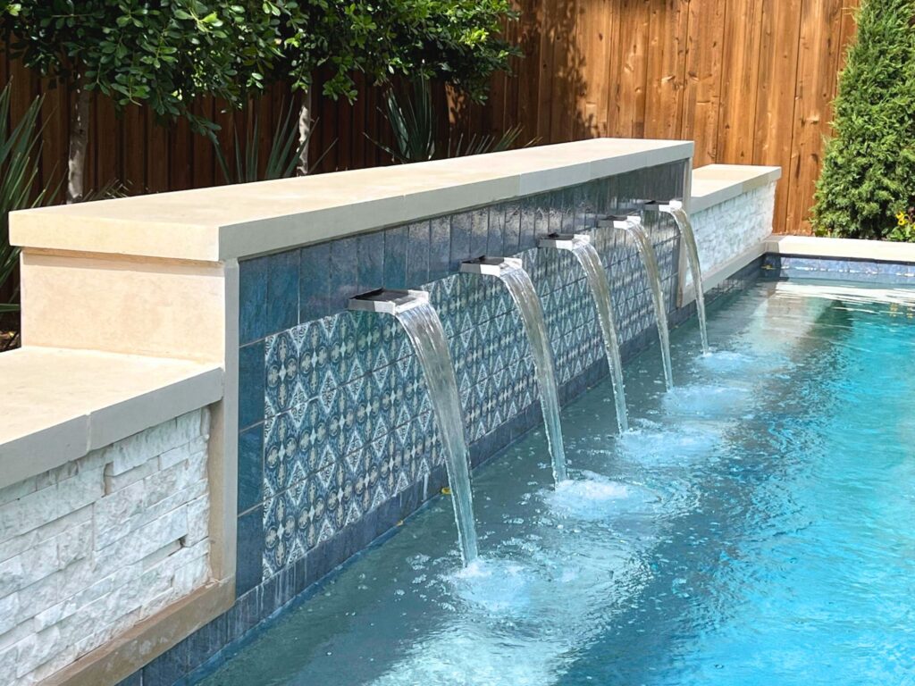 Backyard pool with decorative tile water feature, five spouts flowing into clear water, surrounded by wooden fence and greenery.