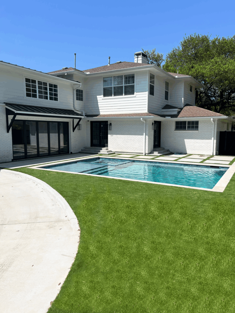 Modern backyard with rectangular pool, shallow entry, concrete deck, green lawn, and two-story white house with black-framed windows and awning