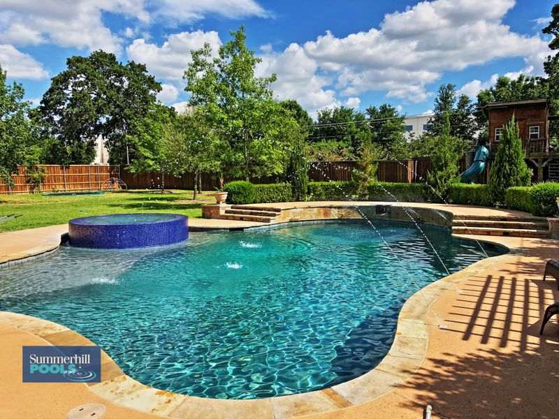 Backyard swimming pool with elevated circular spa, waterfall feature, arching water jets, and lush landscaping including trees and manicured lawn.