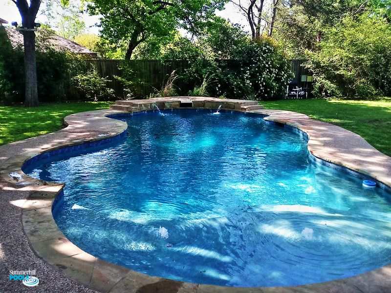 Freeform backyard pool with clear blue water, stone deck, water jets, and lush landscaping including lawn, trees, and shrubs.