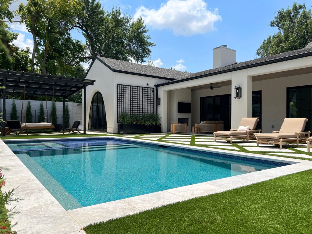 A stunning "after" shot of a luxury backyard transformation, showcasing a rectangular pool with a blue-tiled sun shelf and integrated spa. The design features a striking diamond-patterned patio of white stone and artificial turf, tan lounge chairs, and a modern black pergola with a hanging daybed, all bordered by a row of privacy evergreens.