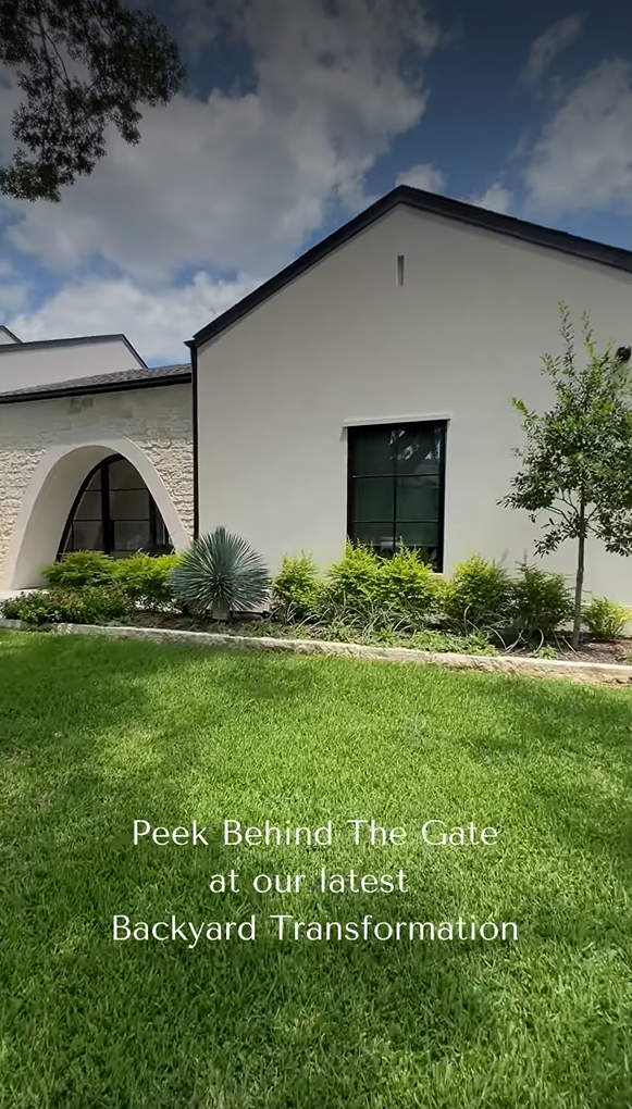 An exterior front view of a modern white stucco home with a green lawn and manicured landscaping. The image features a large arched window with stone accents and black-framed windows, overlaid with the text, "Peek Behind The Gate at our latest Backyard Transformation."
