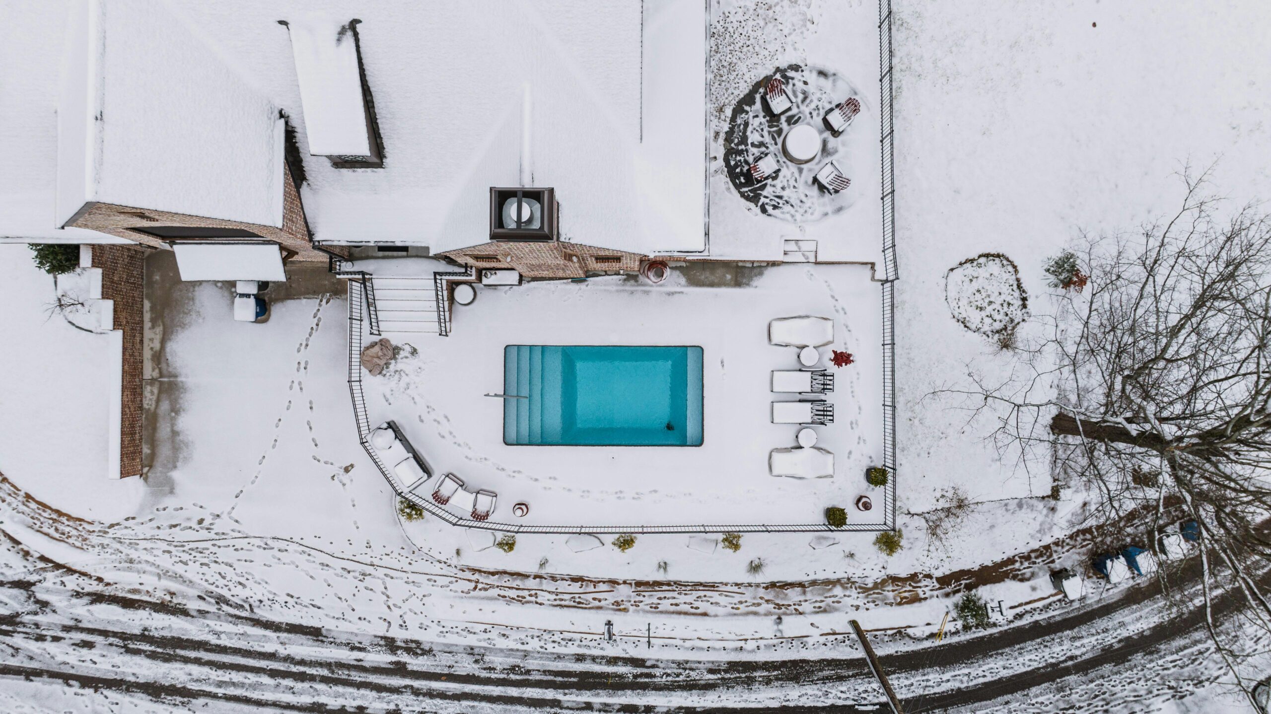 An aerial bird's-eye view of a rectangular backyard pool with bright turquoise water, creating a striking contrast against a snow-covered patio and landscape. The winter scene features a brick home, a fenced-in pool deck with snow-dusted lounge furniture, and a circular fire pit area, demonstrating the year-round aesthetic appeal of a custom pool.
