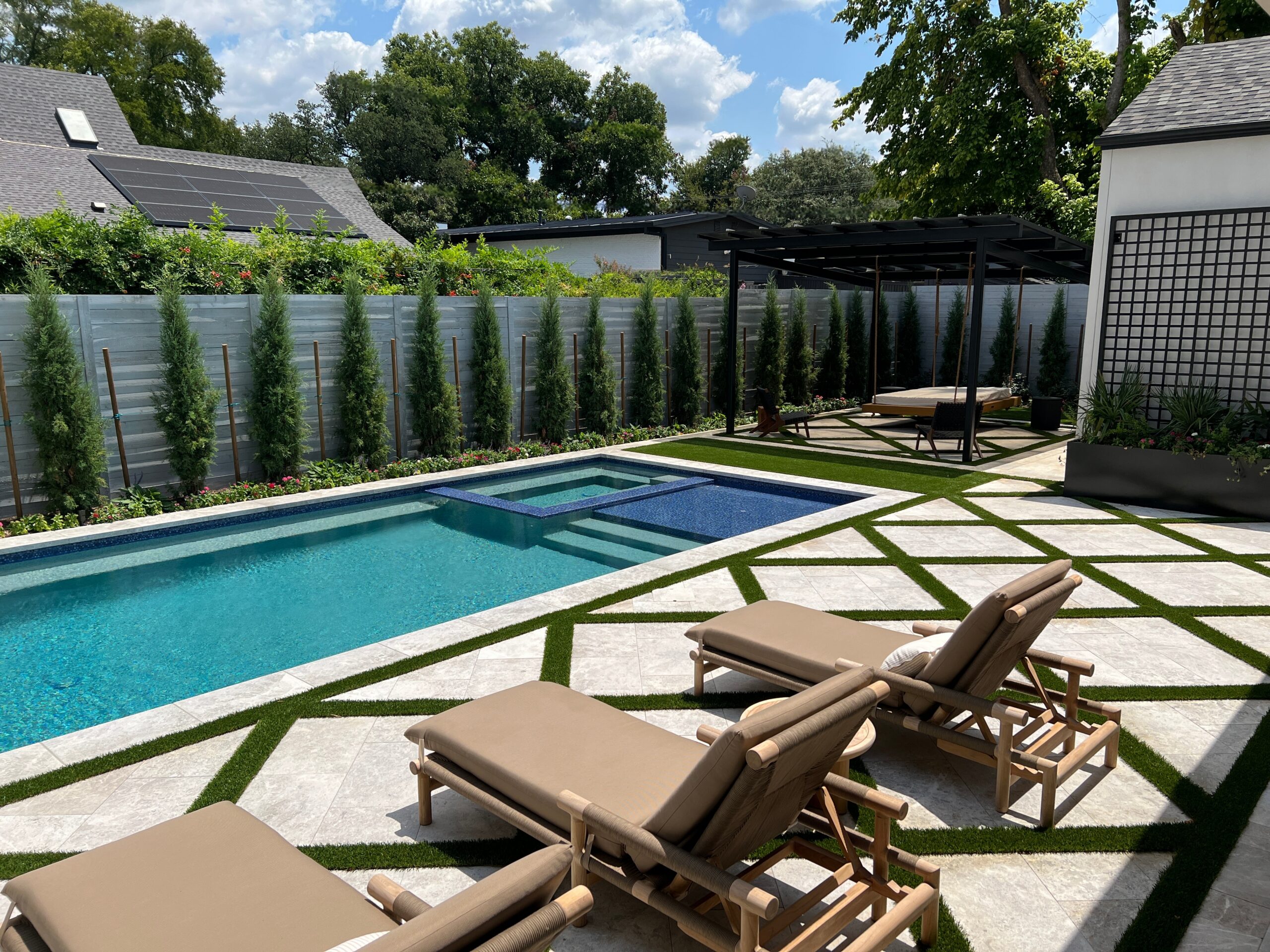 A stunning "after" shot of a luxury backyard transformation, showcasing a rectangular pool with a blue-tiled sun shelf and integrated spa. The design features a striking diamond-patterned patio of white stone and artificial turf, tan lounge chairs, and a modern black pergola with a hanging daybed, all bordered by a row of privacy evergreens.