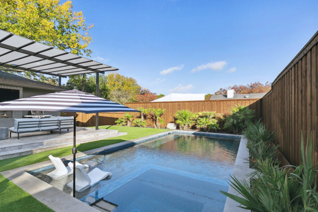 Modern geometric pool design featuring a central square hot tub, arching water fountains, and a tan stone deck with minimalist landscaping.
