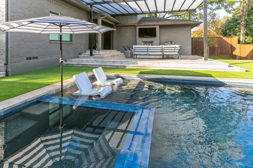 Modern geometric pool design featuring a central square hot tub, arching water fountains, and a tan stone deck with minimalist landscaping.