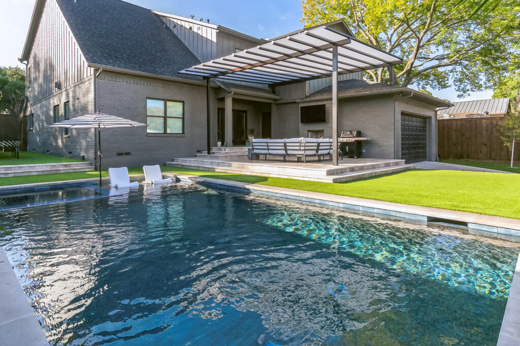 Modern geometric pool design featuring a central square hot tub, arching water fountains, and a tan stone deck with minimalist landscaping.