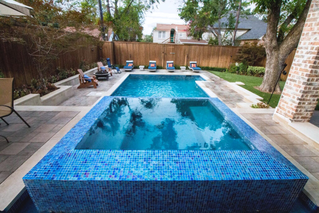 Modern geometric pool design featuring a central square hot tub, arching water fountains, and a tan stone deck with minimalist landscaping.