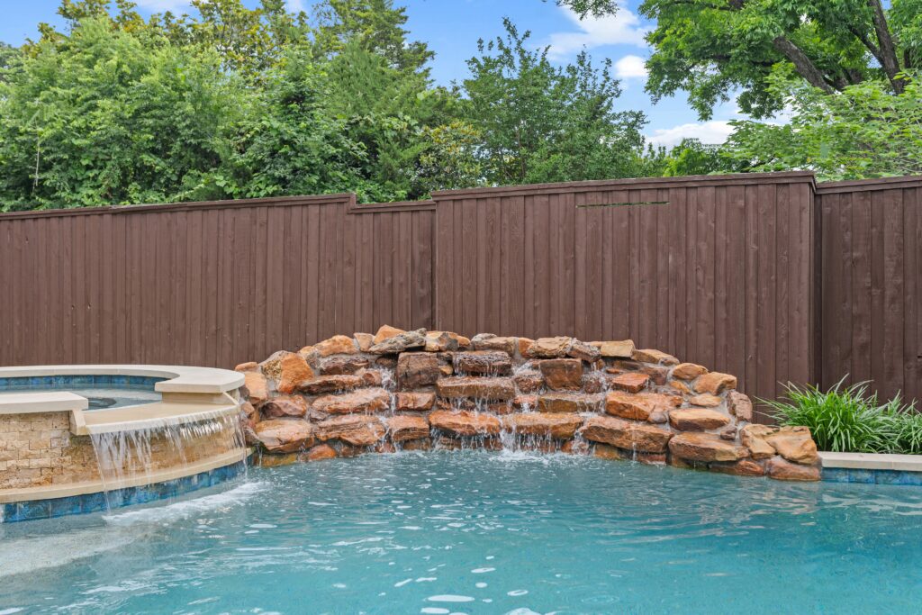 Rustic backyard pool design with a cascading rock water feature, stone-faced spa, and lush perimeter landscaping.