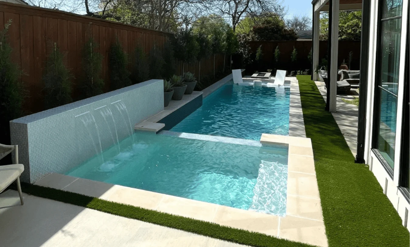 Contemporary pool design featuring a raised light-blue tile wall with waterfalls, white sun shelf loungers, and clean-line stone coping.
