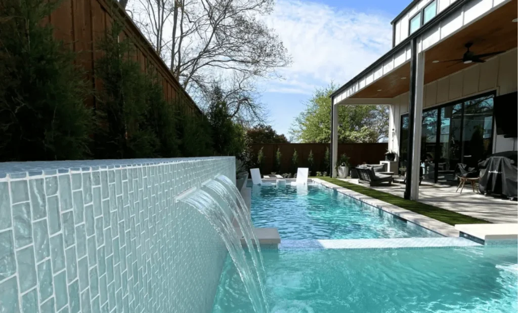 Modern backyard pool with light blue glass tile, integrated three-port water shear feature, artificial turf inlay, and white in-pool lounge chairs.