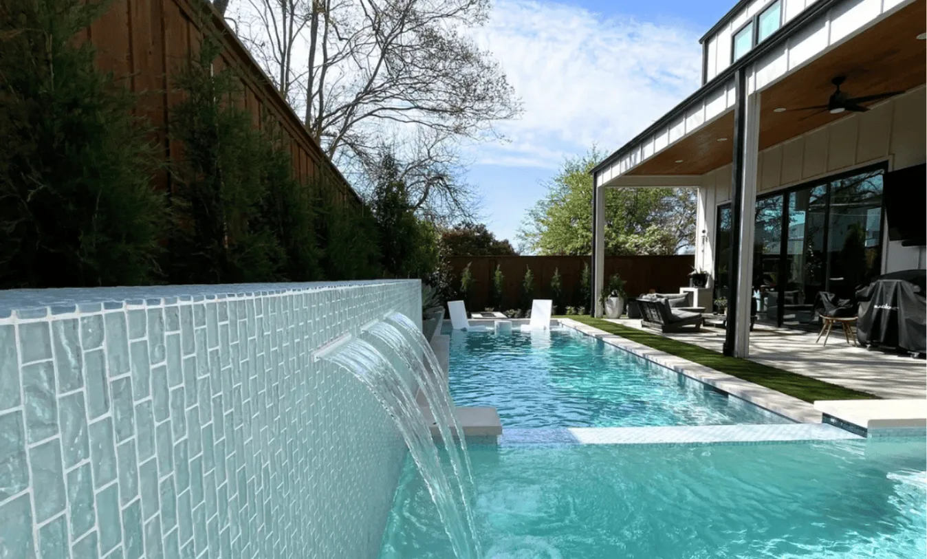 Modern backyard pool with light blue glass tile, integrated three-port water shear feature, artificial turf inlay, and white in-pool lounge chairs.