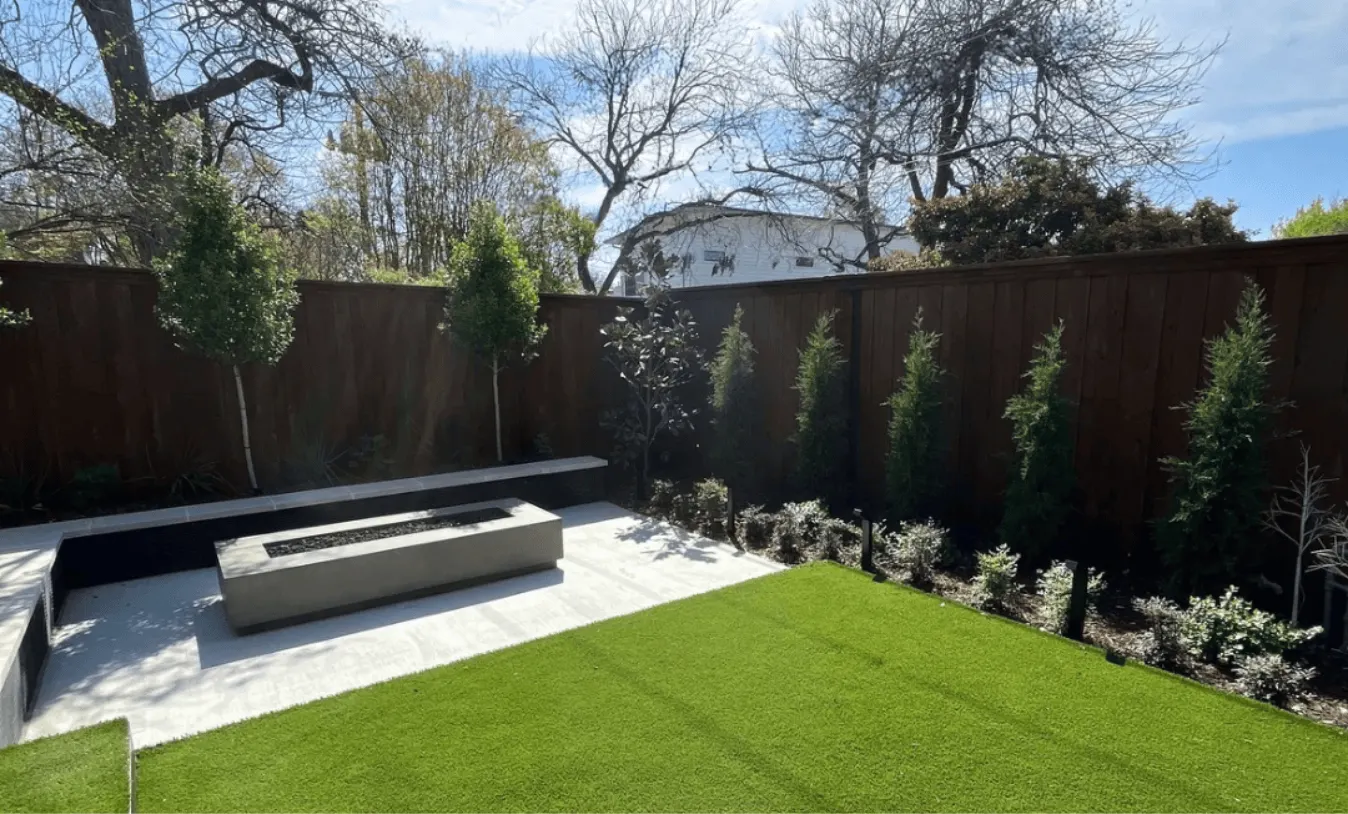 Modern backyard outdoor living space with rectangular concrete gas fire pit, built-in bench seating, large-format concrete pavers, artificial turf lawn, mixed privacy landscaping, and cedar privacy fence in a Dallas backyard