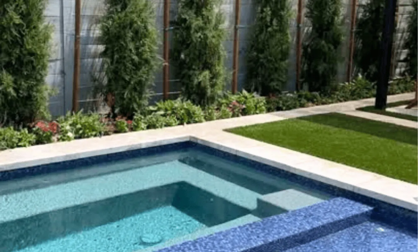 Custom inground pool featuring a built-in sun shelf and bench, surrounded by clean-line stone pavers, artificial turf, and a lush garden border.