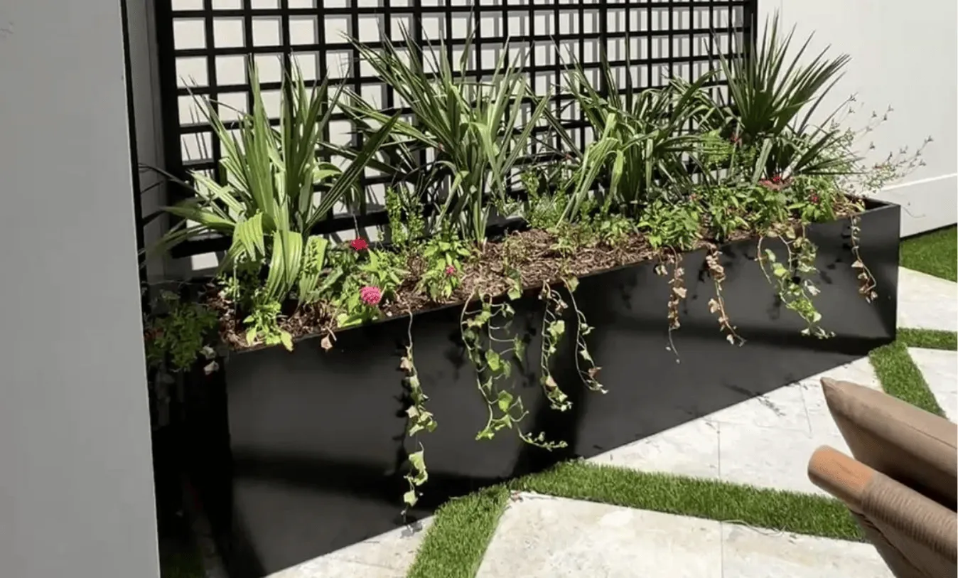 Minimalist black metal garden planter with various green plants on a geometric stone and artificial grass pool deck.