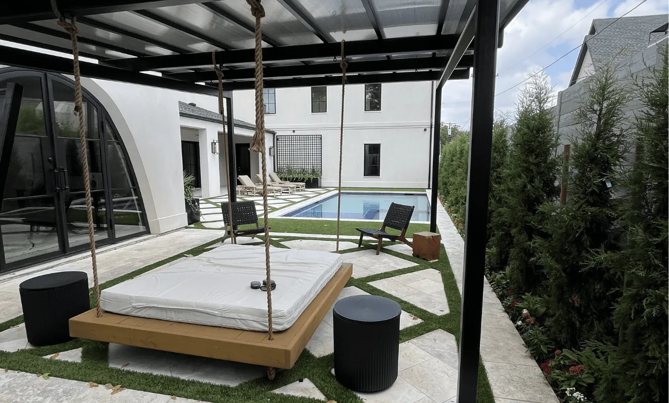 Modern outdoor living space with a large swinging daybed, diamond-patterned paver patio, and an inground lap pool adjacent to a white stucco farmhouse.
