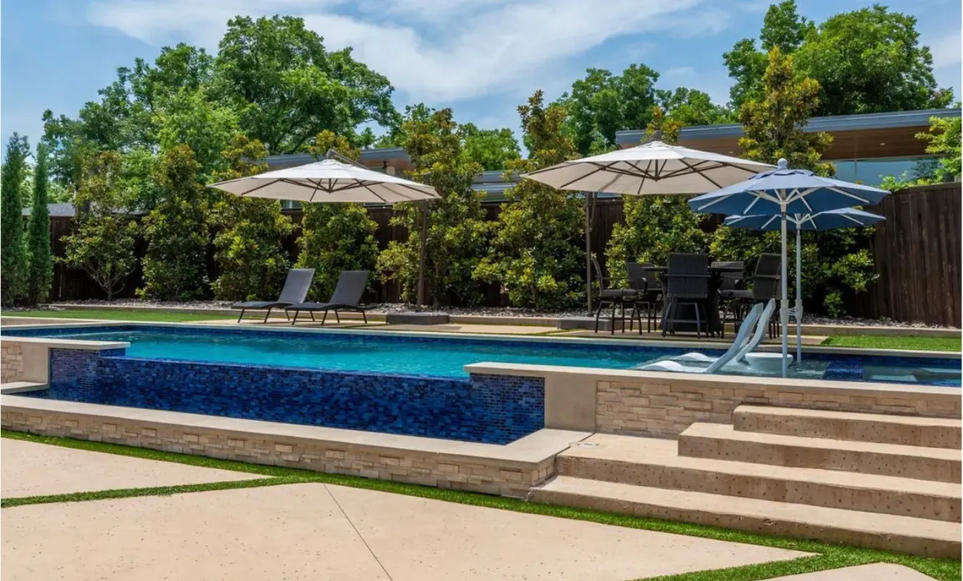 Custom multi-level swimming pool with a raised dark blue mosaic tile water feature, light stone steps, and a sun shelf with umbrellas in Arlington, Texas.