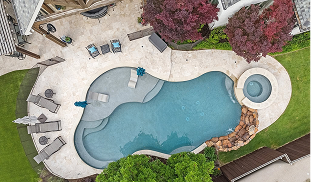 Aerial drone view of freeform inground pool with attached spa, natural stone grotto, travertine deck, lounge chairs, and mature red maple and oak landscaping in a luxury Dallas backyard