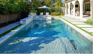 Rectangular lap pool with beach entry, white marble coping, raised spa wall with scupper, travertine deck, artificial turf inlays, cantilever umbrella, and arched covered loggia on Mediterranean luxury Dallas home