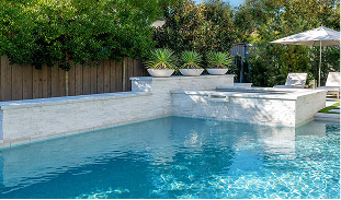 Rectangular inground pool with white marble coping and raised planter wall, decorative bowl planters, scupper waterfall, travertine deck, cantilever umbrella, and mature privacy landscaping