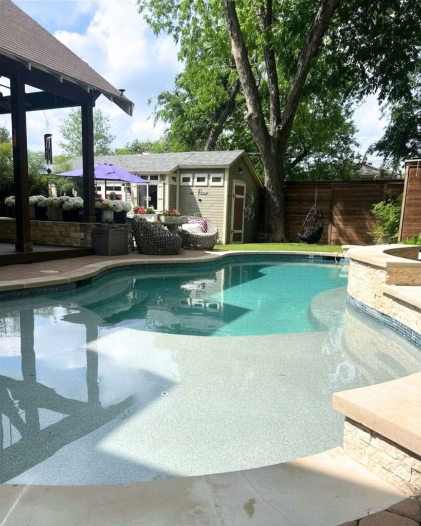 Freeform inground pool with tanning ledge, raised stacked stone spa, travertine coping, covered pergola, pool house, hanging egg chair, artificial turf lawn, and cedar privacy fence in a Dallas backyard