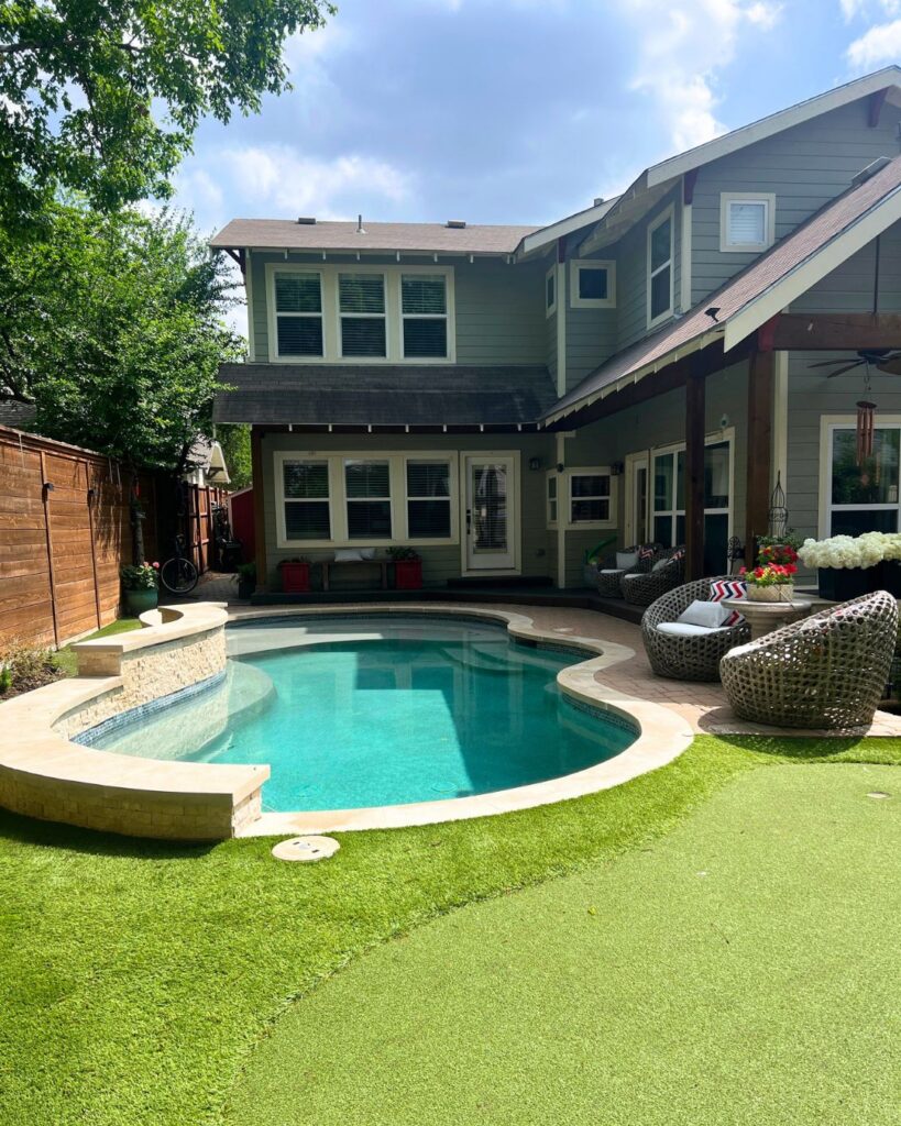 Freeform inground pool with tanning ledge, raised stacked stone spa, travertine coping, covered pergola, pool house, hanging egg chair, artificial turf lawn, and cedar privacy fence in a Dallas backyard