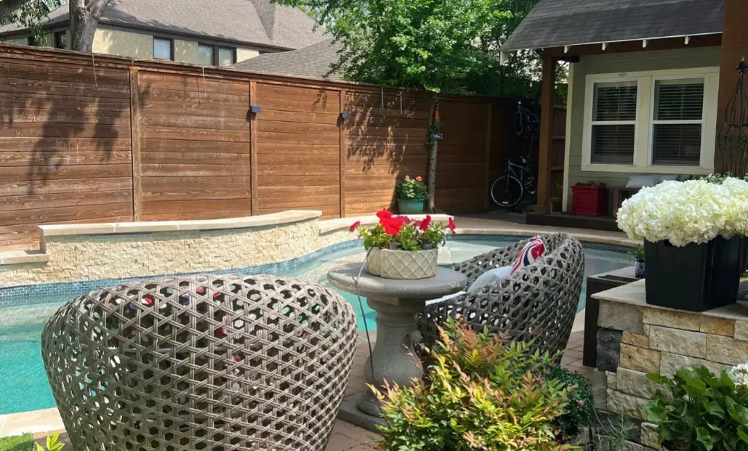 Freeform inground pool with travertine coping, blue glass tile waterline, attached spa, covered pergola, and lush cottage garden patio with potted flowers and cedar privacy fence