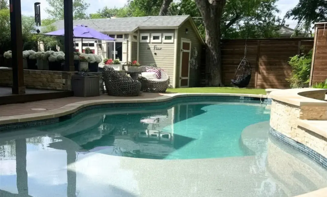 Freeform inground pool with tanning ledge, raised stacked stone spa, IPE wood deck, wicker lounge furniture, hanging egg chair, pool house, artificial turf lawn, and cedar privacy fence in a Dallas backyard