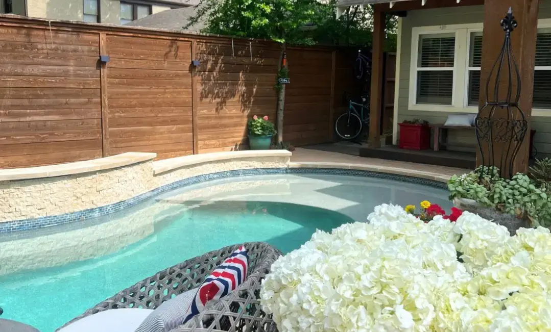 Freeform inground pool with tanning ledge, raised stacked stone spa, IPE wood deck, wicker lounge furniture, hanging egg chair, pool house, artificial turf lawn, and cedar privacy fence in a Dallas backyard