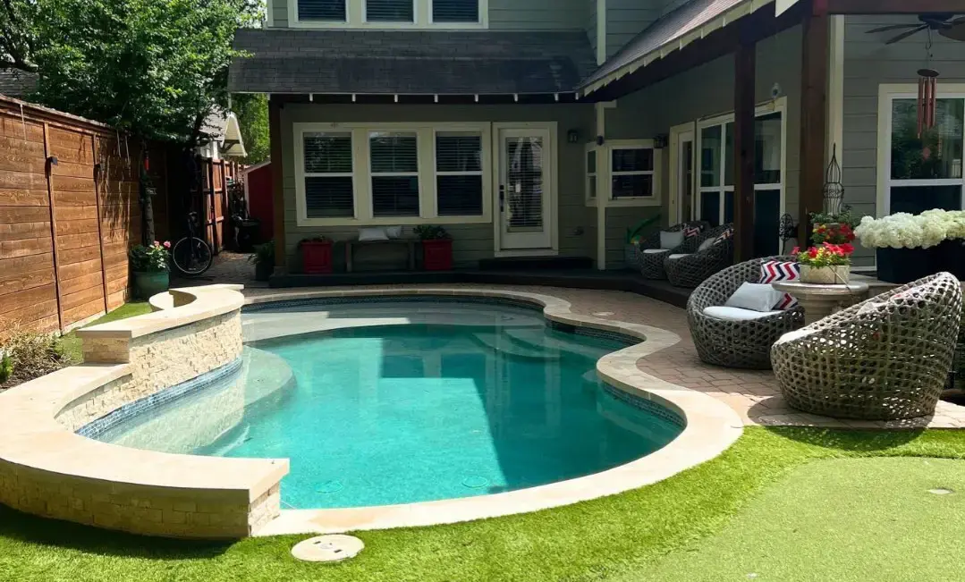 Freeform inground pool with tanning ledge, raised stacked stone spa, IPE wood deck, wicker lounge furniture, hanging egg chair, pool house, artificial turf lawn, and cedar privacy fence in a Dallas backyard