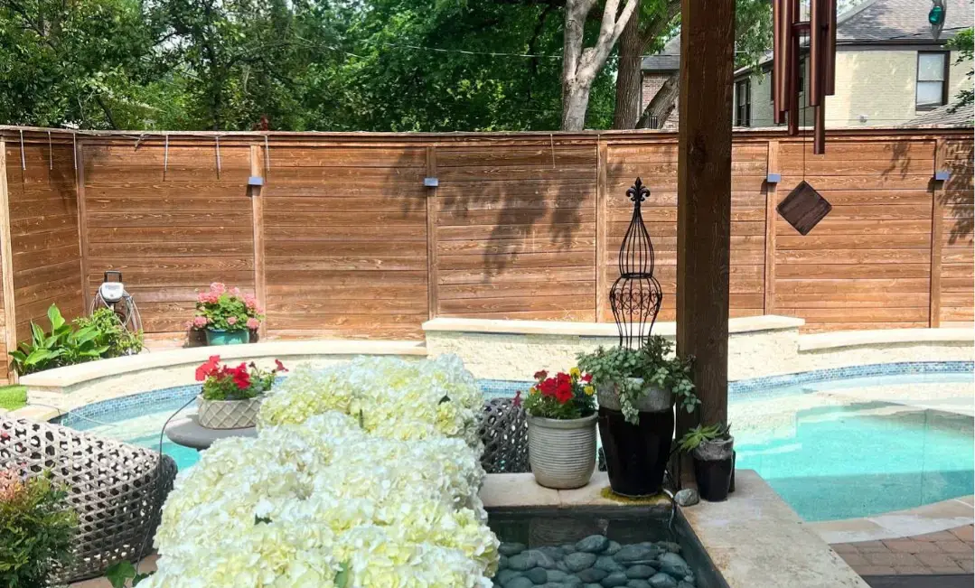 Freeform inground pool with travertine coping, blue glass tile waterline, attached spa, covered pergola, and lush cottage garden patio with potted flowers and cedar privacy fence