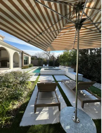 A luxurious backyard pool and lounging area viewed from beneath a tan and white striped patio umbrella. The scene showcases two wooden lounge chairs on a geometric white stone and turf patio, leading toward a modern rectangular pool with an integrated raised stone spa and a Mediterranean-style outdoor kitchen in the background.