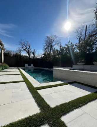 An ultra-modern backyard pool featuring white stone pavers with geometric artificial turf inlays. The rectangular swimming pool has dark teal water, complemented by a raised stone spa and a clean, minimalist patio design under a bright, clear blue sky.