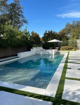 High-angle wide shot of a completed luxury pool and spa in Dallas, TX, featuring white stone decking with turf inlays, contemporary white loungers, and tiered masonry water features.