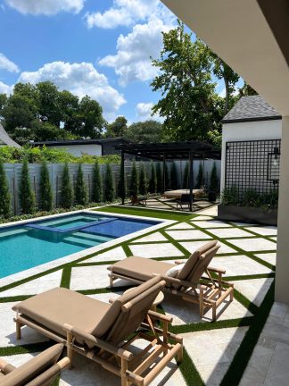 A wide-angle view of a luxury backyard featuring a geometric stone and turf patio next to a swimming pool. Modern lounge chairs sit in the shaded foreground, while a black trellis, planter, and row of cypress trees line the perimeter under a bright sky.