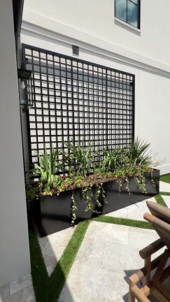 A black rectangular planter box filled with green leafy plants and vines sits against a white exterior wall. Behind the plants, a tall black metal grid trellis is mounted to the wall, creating a modern architectural accent above a stone and turf patio.