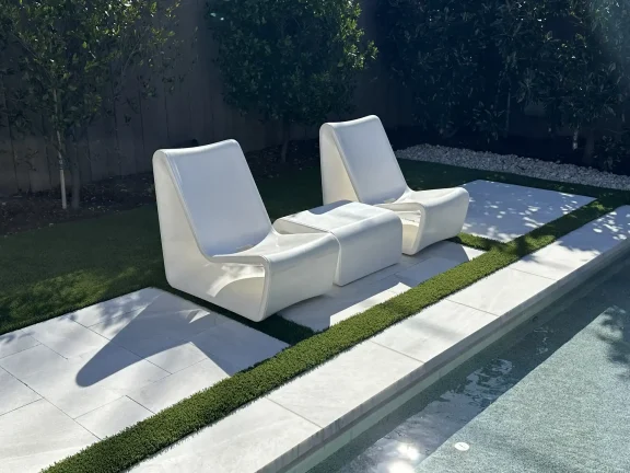 Elegant white Ledge Loungers and side table set on a travertine and turf patio in Arlington, TX, overlooking a modern geometric pool.