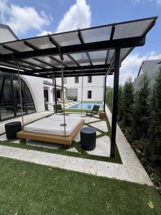A modern backyard features a wooden hanging daybed suspended by ropes under a black pergola. The daybed sits on a geometric stone and turf patio next to a swimming pool. In the background, a white house with large arched windows and a row of evergreen trees complete the luxury outdoor space.