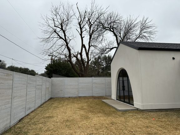 A "before" shot of a flat, grassy backyard featuring a modern white guest house with a large arched glass door. The space is enclosed by a light gray horizontal wood fence, providing a blank canvas for a custom pool and landscape design under an overcast sky.