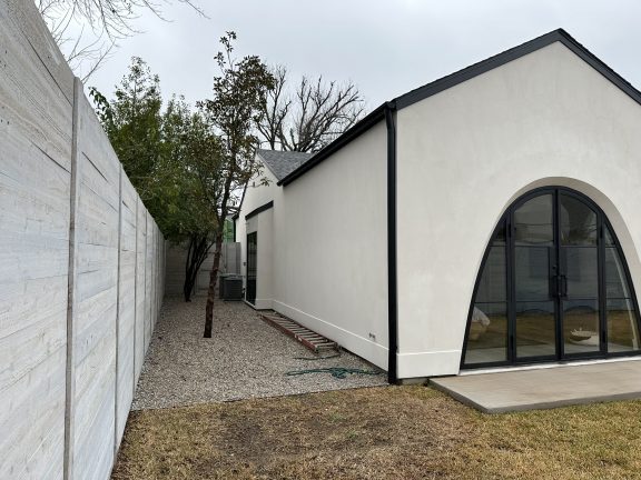A white modern gabled building with large arched black-trimmed glass doors. A narrow side yard with gravel, a small tree, and a ladder sits between the building and a light gray wooden fence under an overcast sky.