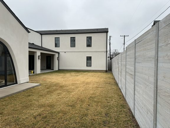 A "before" shot of a flat, grassy backyard featuring a modern white guest house with a large arched glass door. The space is enclosed by a light gray horizontal wood fence, providing a blank canvas for a custom pool and landscape design under an overcast sky.